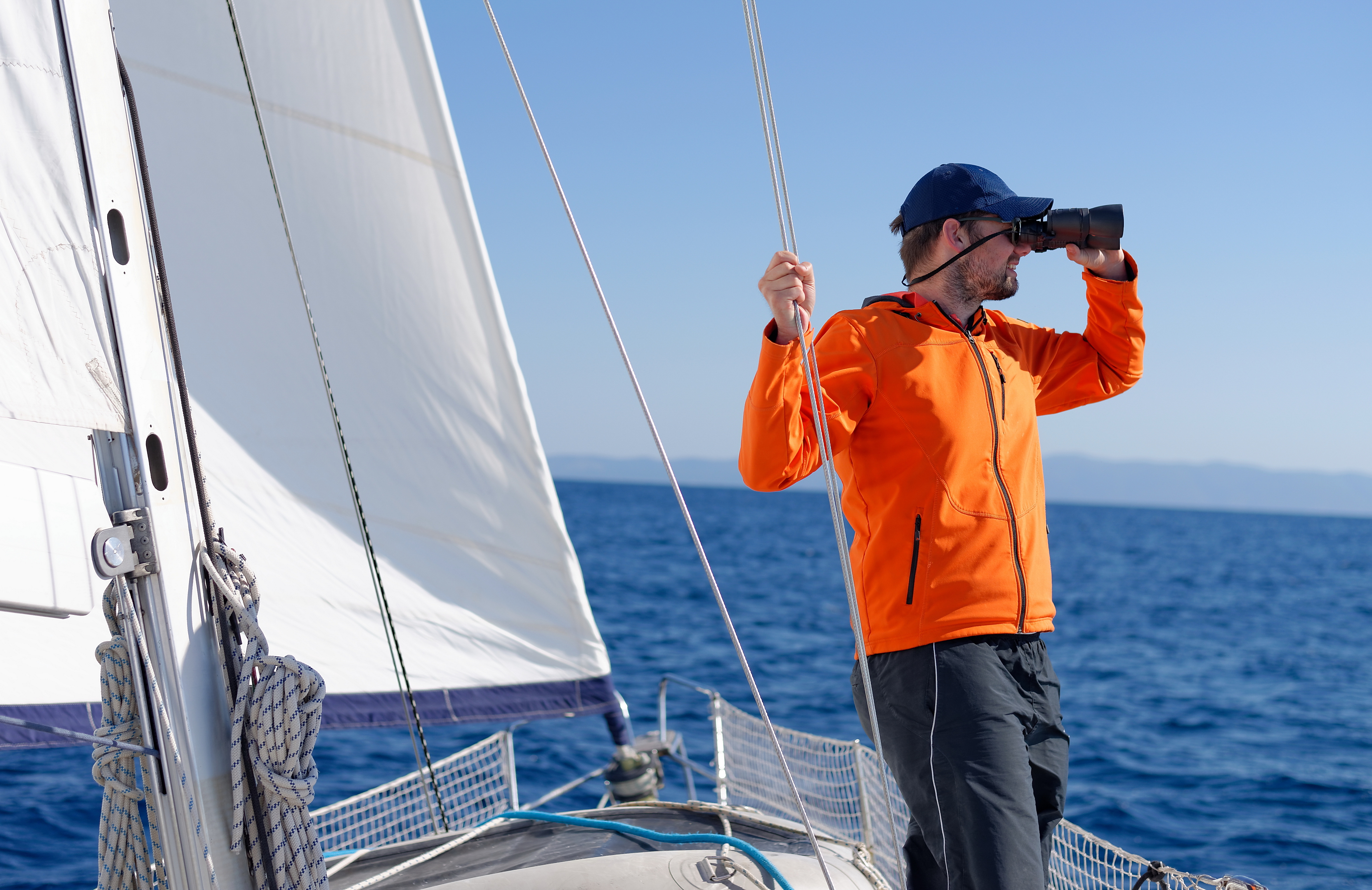 A man in an orange jacket using binoculars on a sailboat. 
