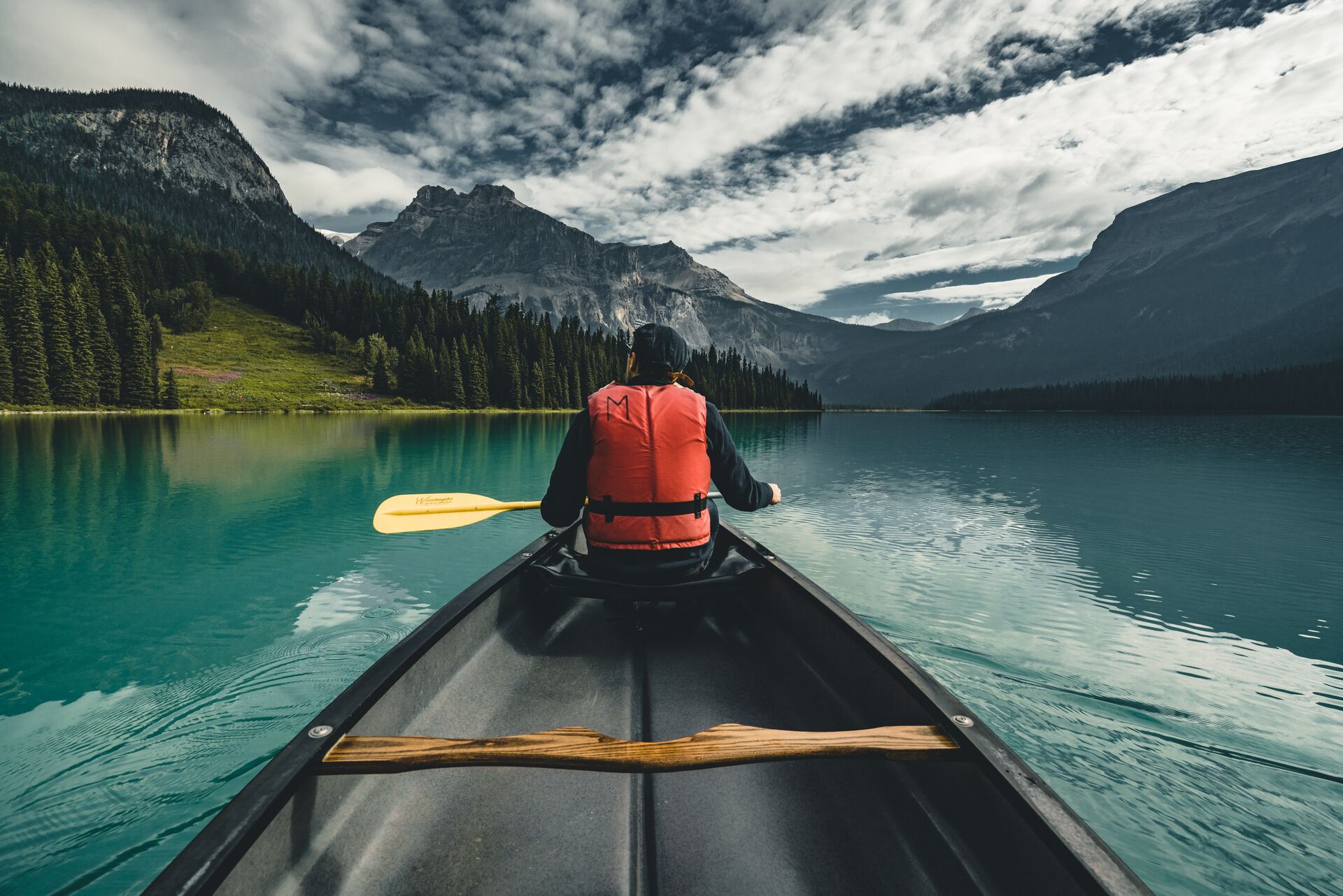 A man sitting at the front of a canoe on the water with a paddle looking at mountains. 