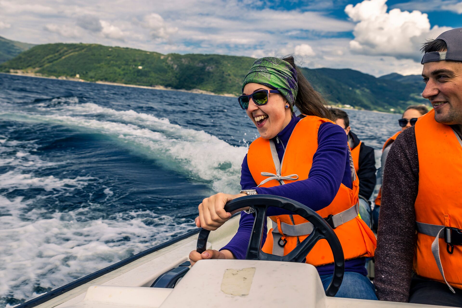 A smiling woman in an orange life jacket drives a boat. 