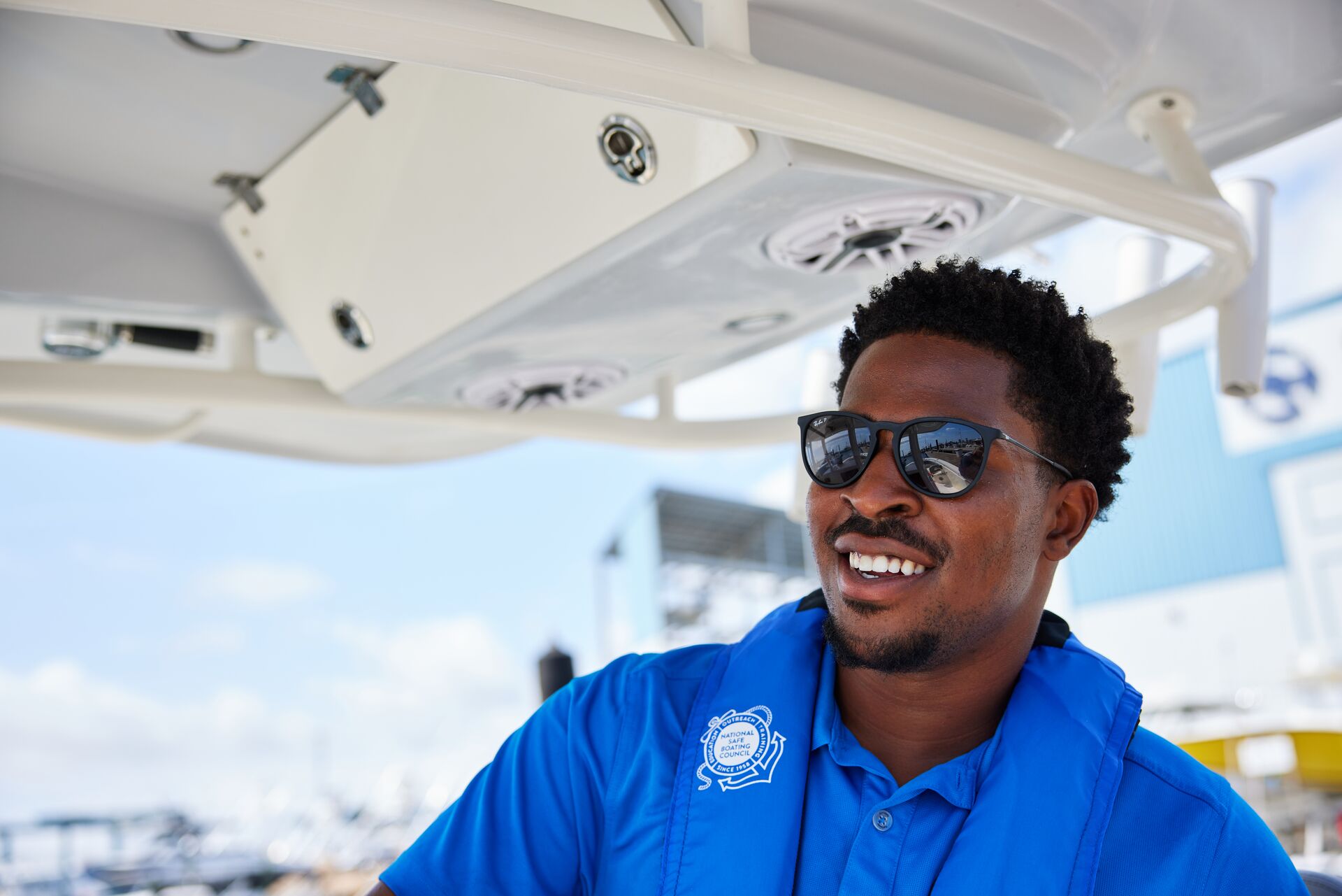 A man in a blue life jacket smiling while driving a boat. 